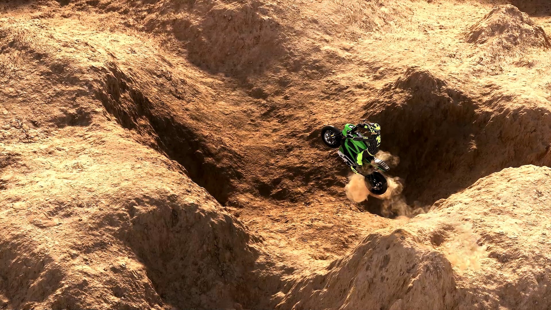 A rider on a green motorcycle in a sandy desert canyon, seen from above.